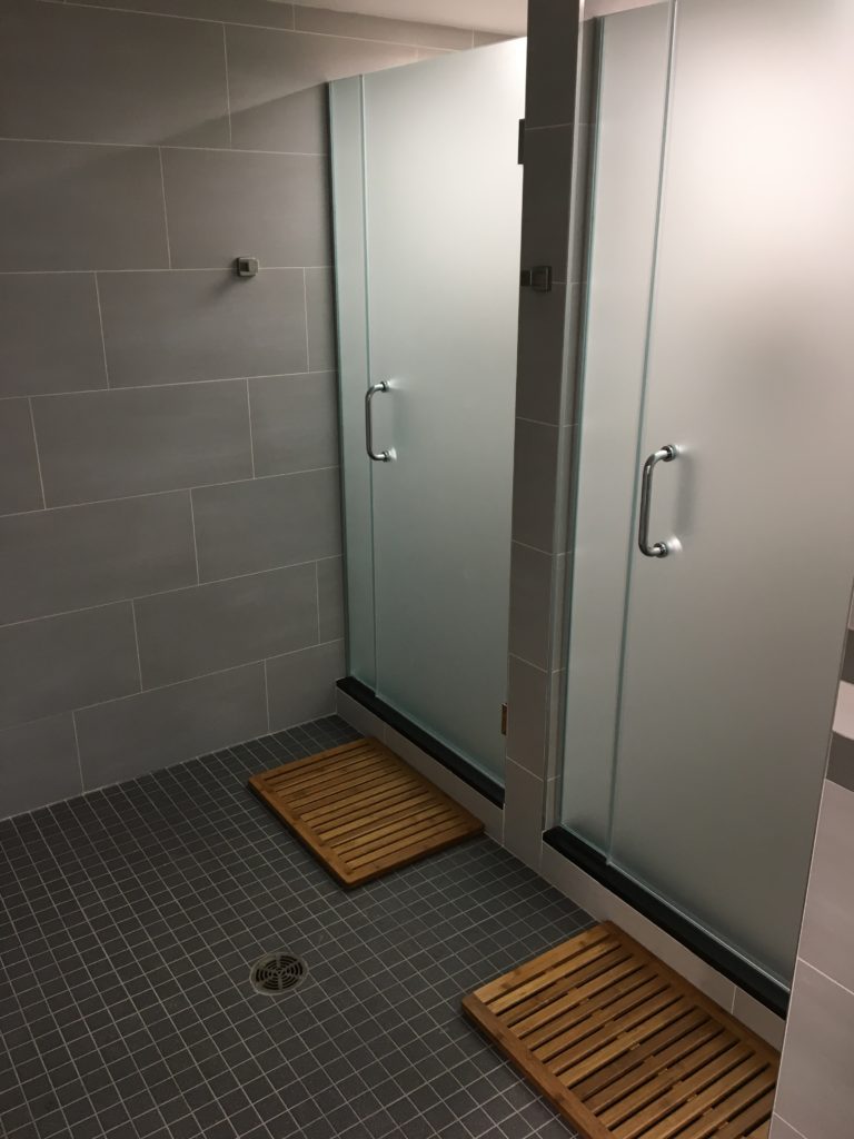 Private showers with frosted glass doors, gray stone tile, and teak wood mats